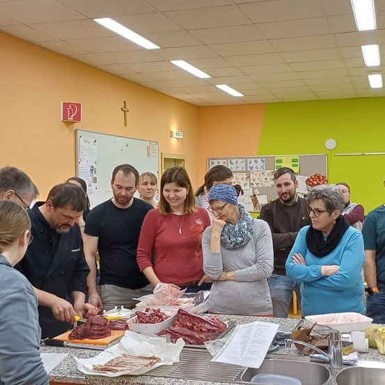 Mehrere Personen versammeln sich um einen Küchentresen, wo eine Person Fleisch schneidet. Verschiedene Fleischsorten liegen auf Tabletts und Schneidebrettern. Die Frau in der Mitte trägt einen Schal und hat ihre Hand am Kinn. Hinter ihnen hängen eine Tafel und ein Kreuz an der Wand.