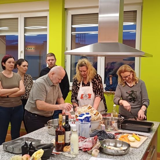 Eine Gruppe von Menschen, darunter Männer und Frauen, steht um einen Küchentisch herum und bereitet Essen zu. Die Küche hat große Fenster und eine gelbe Wand.