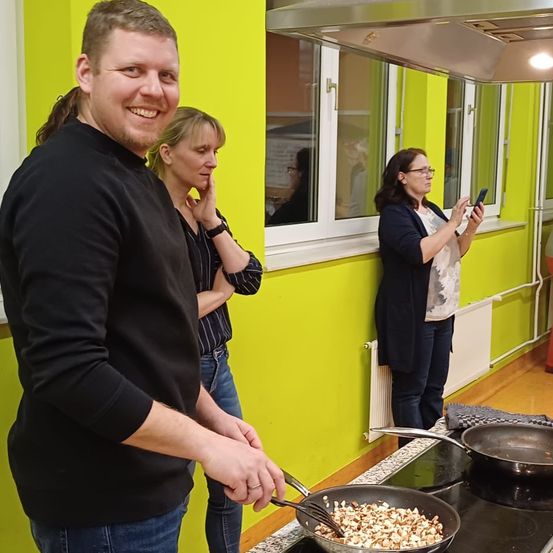 Ein Mann in einem schwarzen Shirt kocht am Herd. Zwei Frauen beobachten ihn, eine mit einem Telefon. Die Küche hat gelbe Wände und Fenster.