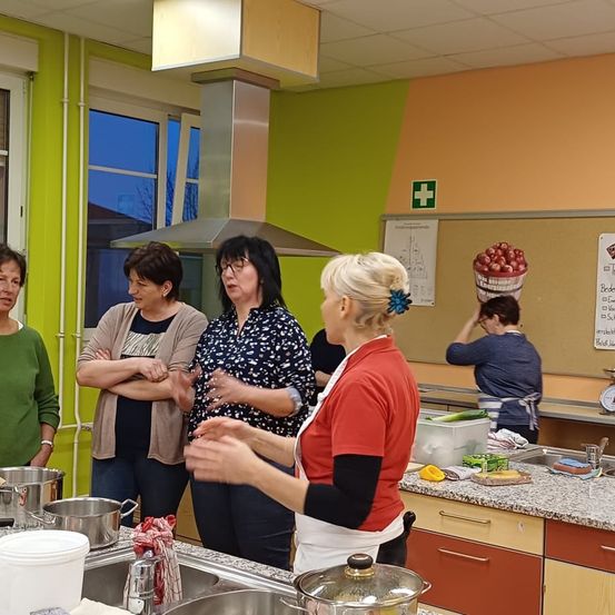 Eine Gruppe von Frauen befindet sich in einer Küche, einige stehen und andere sind in der Nähe des Waschbeckens. Sie scheinen zu sprechen. Töpfe, Pfannen und Schüsseln stehen auf der Arbeitsplatte. Eine Tafel mit einem Bild von Äpfeln ist an der Wand.