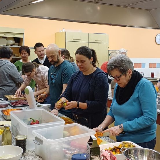 Eine Gruppe von Menschen kocht in einer Küche, wobei mehrere Erwachsene mit der Essenszubereitung beschäftigt sind. Sie verwenden Plastikbehälter und ein Schneidebrett. Eine Frau im Vordergrund schneidet Gemüse.