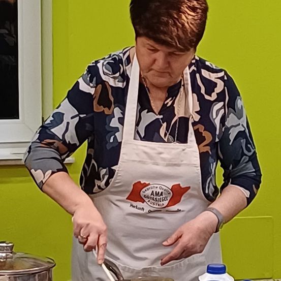 Eine Frau mit ernster Miene rührt in einer Küche einen Topf. Sie trägt ein Blumenhemd und eine weiße Schürze mit einem Emblem. Eine grüne Wand und ein Fenster befinden sich hinter ihr.