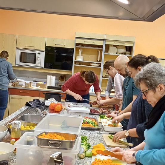 Eine Gruppe von Menschen in einer Küche bereitet Essen zu, wobei eine Frau ein rotes Shirt trägt und andere verschiedene farbige Oberteile. Küchengeräte und verschiedene Lebensmittel sind auf dem Tresen.
