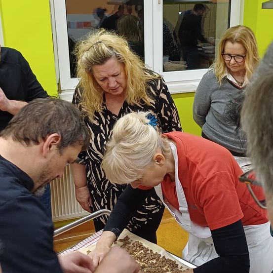 Mehrere Personen sind in einer Küche. Eine Frau in einem roten Shirt beugt sich über eine Schale, und ein Mann in einem blauen Shirt schaut zu ihr. Dahinter steht eine Frau in einem schwarz-weißen Kleid neben einem Glasfenster.