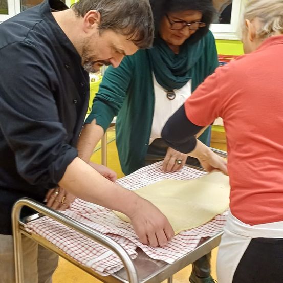 Drei Personen bereiten gemeinsam einen Teig auf einem Tisch vor, der Mann in Schwarz und die Frau in Grün beobachten den Teig aufmerksam, während die Frau in Rot darauf zeigt.