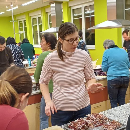 Eine Gruppe von Menschen ist in einer Küche und bereitet Essen zu. Eine Frau in einem gestreiften Shirt erklärt etwas den anderen, während eine Schale mit Essen auf Stöcken auf dem Tresen steht.