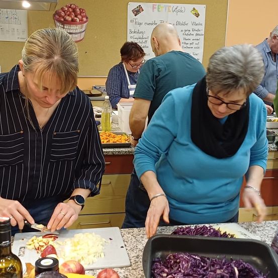 Mehrere Personen befinden sich in einer Küche und bereiten Essen zu. Eine Frau schneidet Äpfel auf einem Schneidebrett. Eine andere Frau rührt etwas in einer Schüssel. Ein Mann steht hinter ihnen.