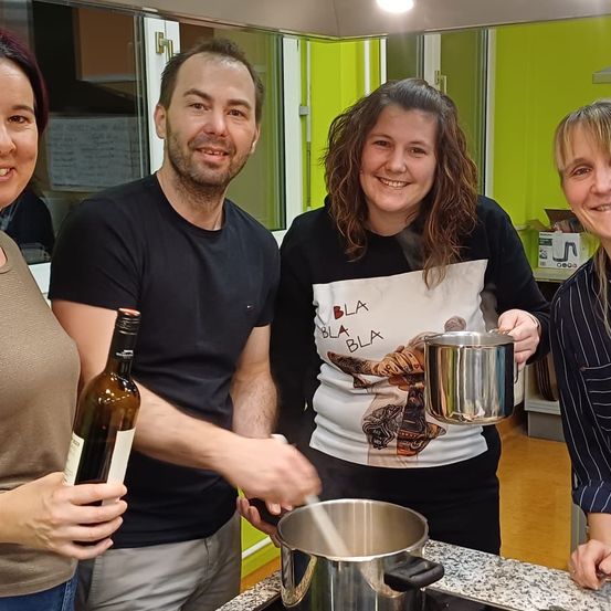 Vier Menschen stehen in einer Küche mit grünen Wänden. Sie lächeln und scheinen zu kochen. Ein Mann hält eine Flasche Wein, während eine Frau einen silbernen Topf hält. Eine weitere Frau hält einen Topf und eine Flasche. Die vierte Person steht ganz rechts.