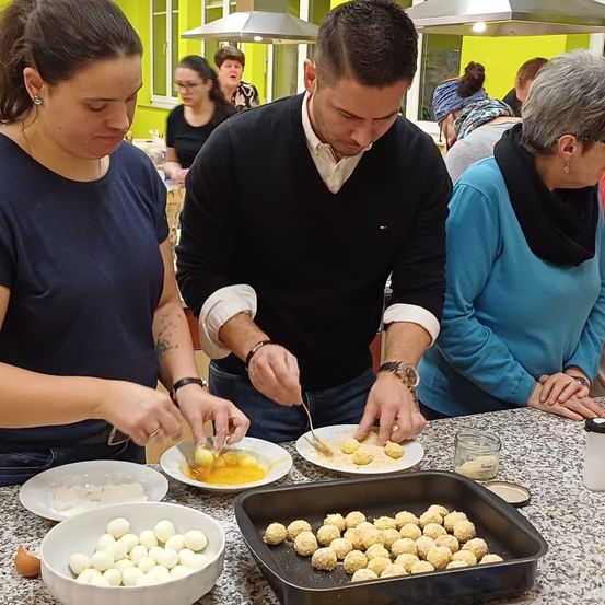 Eine Gruppe von Menschen kocht in einer Küche. Ein Mann und eine Frau mischen Zutaten. Sie haben Teller und Schüsseln mit Essen. Andere Menschen beobachten sie.