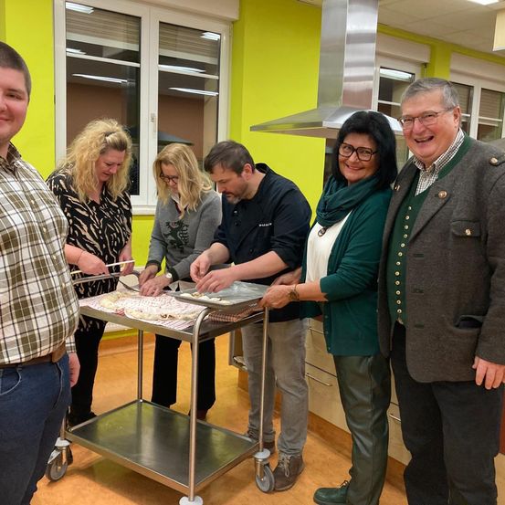 Sechs Personen befinden sich in einer Küche, wobei vier Personen Essen auf einem Wagen zubereiten. Eine Frau hält eine Tablett mit Essen. Der Mann neben ihr trägt einen Pullover und eine Brille. Eine weitere Frau lächelt neben einem Mann in einem karierten Hemd.