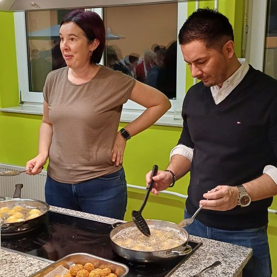 Eine Frau und ein Mann kochen in einer Küche mit grünen Wänden. Die Frau hält einen Kochlöffel, und der Mann benutzt einen Spatel. Ein Glas und eine Uhr sind in der Nähe.