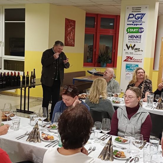 Eine Gruppe von Menschen sitzt an einem Tisch und genießt eine Mahlzeit. Ein Mann steht neben einem Tisch mit Weinflaschen. An der Wand hängen Poster und auf der Fensterbank steht eine Topfpflanze.