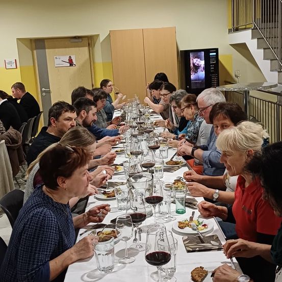 Eine Gruppe von Menschen sitzt an einem langen Tisch in einem Restaurant und genießt eine Mahlzeit mit Wein. Der Tisch ist mit Tellern, Gläsern und verschiedenen Gerichten geschmückt. Einige unterhalten sich, während andere ihr Essen genießen. Der Raum hat eine gelbe Wand mit einem Schild und einer Treppe.