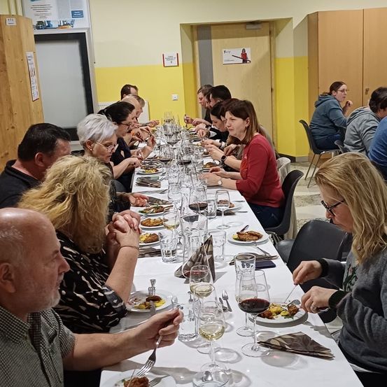Eine Gruppe von Menschen sitzt an einem langen Tisch in einem Raum, isst Essen und trinkt Wein. Auf dem Tisch stehen mehrere Gläser und Teller.