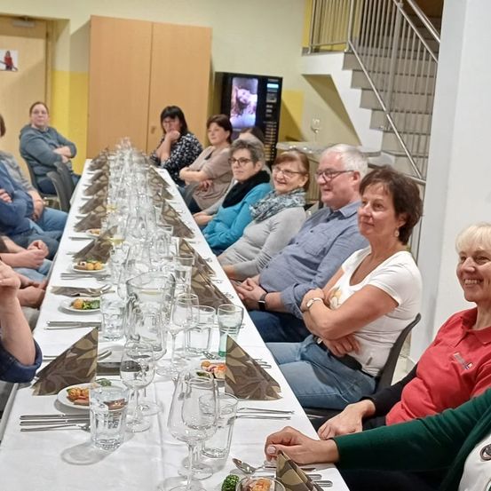 Eine Gruppe von Menschen sitzt an einem langen Tisch in einem Restaurant. Sie lächeln und scheinen ihre Mahlzeit zu genießen. Der Tisch ist mit Tellern, Weingläsern und Servietten gedeckt. Dahinter befindet sich eine Treppe und ein Monitor, der ein Bild anzeigt.
