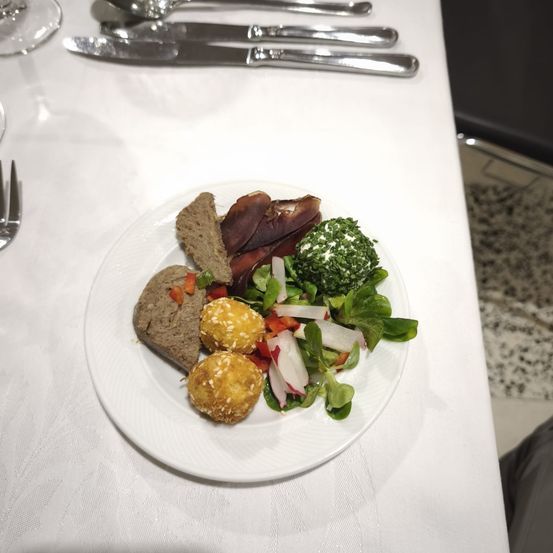 Ein weißer Teller auf einem Tisch enthält verschiedene Speisen: Fleischscheiben, Brot, grünes Gemüse und einen Salat mit roten und weißen Toppings. Silberbesteck steht hinter dem Teller.