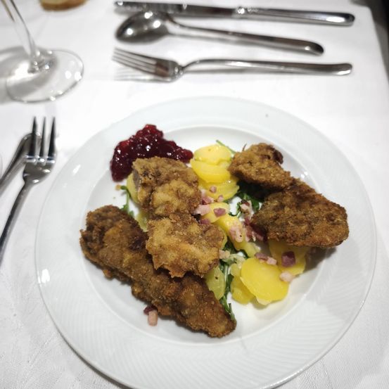 Ein Teller mit gebratenem Fleisch, Kartoffeln und Cranberry-Sauce, begleitet von Besteck und einem Weinglas auf einer weißen Tischdecke.