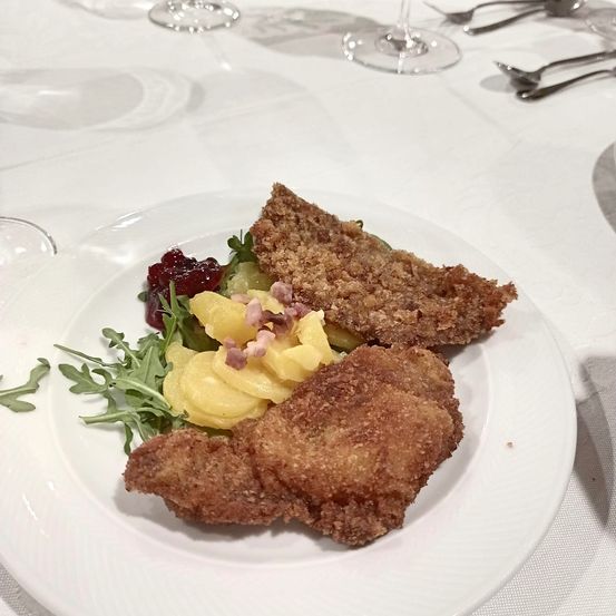 Ein weißer Teller mit gebratenem Fleisch, Kartoffelpüree und Salatgarnitur. Auf einer weißen Tischdecke mit Besteck gedeckt.