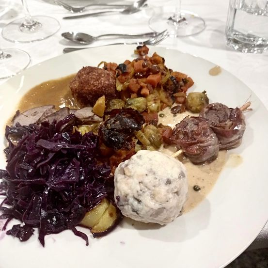 Ein Teller mit verschiedenen Speisen: Fleischbällchen, gebratenes Gemüse und Fleischspieße, begleitet von Rotkohl und einer Knödel.