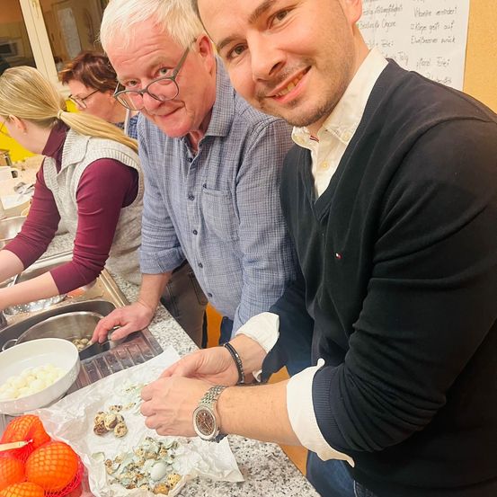 Zwei Männer stehen in einer Küche mit anderen und bereiten Essen zu. Einer schneidet, während der andere lächelt und zeigt. Es gibt Eier, Orangen und eine Schüssel auf dem Tresen.