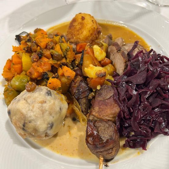 Ein Teller mit verschiedenen Gerichten, darunter ein Fleischspieß, eine Knödel, Rotkohl und eine Mischung aus Gemüse.
