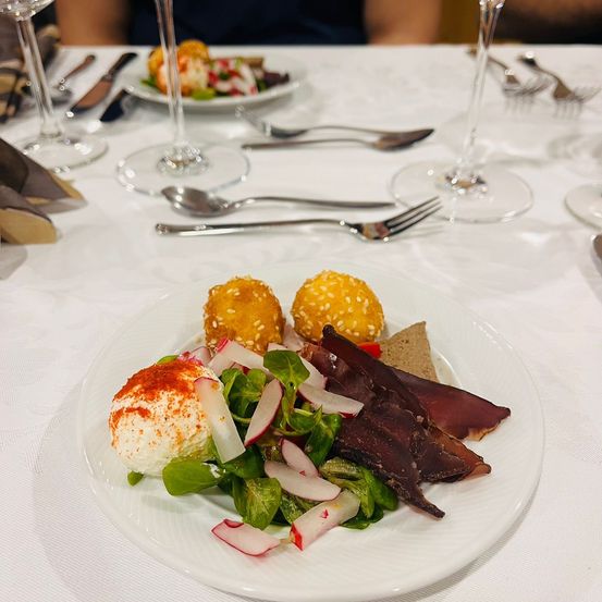 Ein weißer Teller auf einem Tisch mit einem Salat mit Radieschen und getrocknetem Fleisch, zwei frittierten Bällen und einem Käseball mit roten Gewürzen.