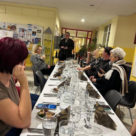 Eine Gruppe von Menschen sitzt an einem langen Tisch, wahrscheinlich in einem Restaurant. Ein Mann steht am Kopf des Tisches und spricht zu der Gruppe. Alle scheinen in ein Gespräch vertieft zu sein. Der Tisch ist mit Tellern, Gläsern und Besteck gedeckt. An der Wand befinden sich Pflanzen und Poster.