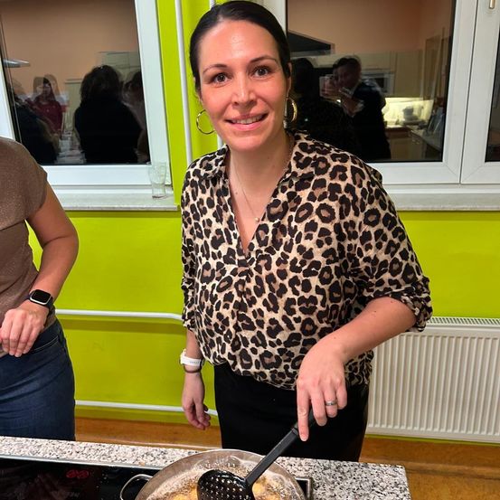 Eine Frau in einem Leopardenmuster-Blusen und schwarzen Hosen kocht in einer Küche. Sie hält einen Schöpflöffel und lächelt. Eine andere Person steht hinter ihr, und ein Küchenfenster zeigt die Reflexion von Menschen.