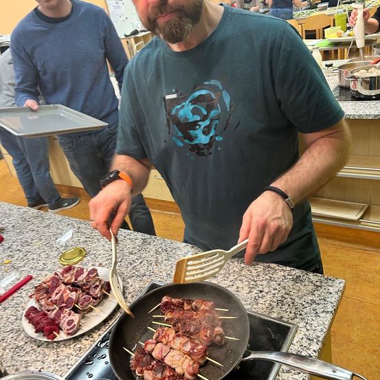 Ein Mann in einem grünen T-Shirt kocht Fleisch in einer Pfanne auf dem Herd. Ein Teller mit Fleisch steht auf der Theke. Ein weiterer Mann steht hinter ihm und hält eine Schale.