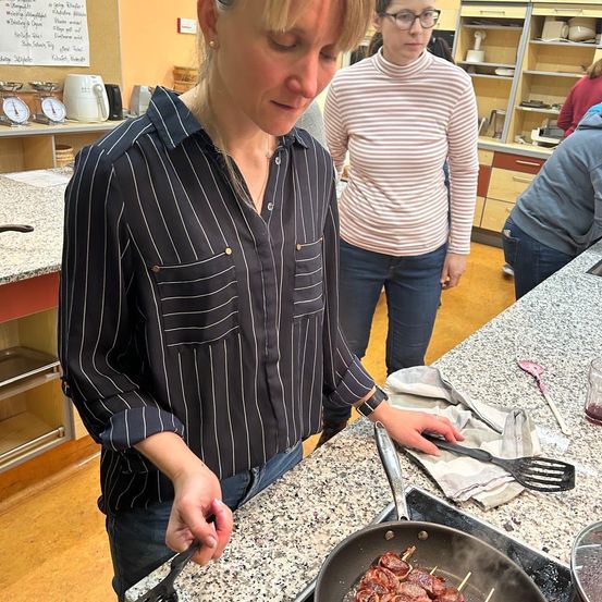 Eine Frau kocht in einer Küche auf einem Herd, hält eine Pfannenwender und schaut nach unten. Eine andere Frau steht in der Nähe und beobachtet. Dahinter befindet sich eine Küche mit Schränken, Regalen und einem weißen Kühlschrank.