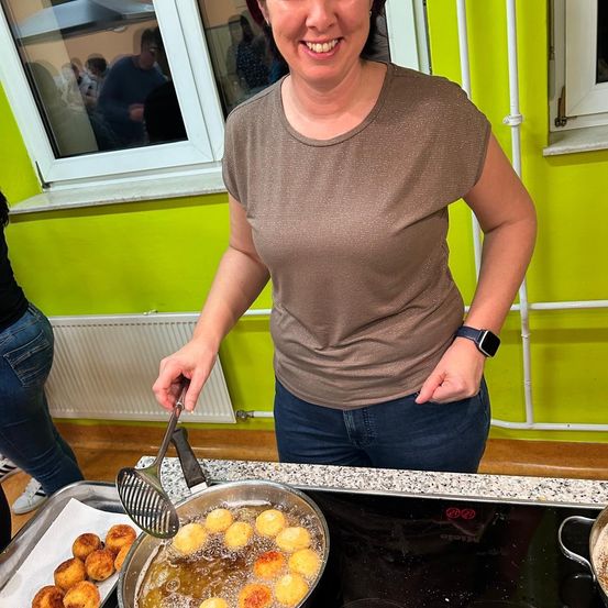 Eine Frau kocht Essen auf einem Herd. Sie lächelt und hält einen Schöpflöffel. Es gibt Teller mit Essen.