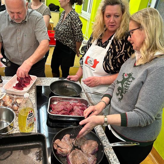 Eine Gruppe von Menschen befindet sich in einer Küche und bereitet Essen zu. Ein älterer Mann schneidet Fleisch, und eine Frau benutzt einen Kochlöffel. Eine andere Frau schaut sich das geschnittene Fleisch an. Hinter ihnen steht ein Herd mit einer Pfanne mit Fleisch und einer Flasche Öl.