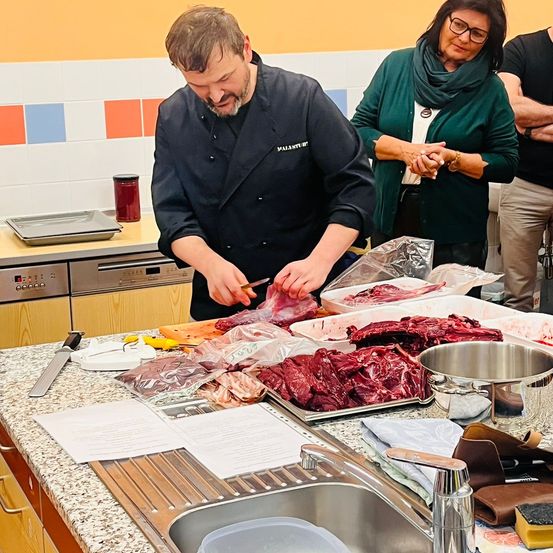 Ein Koch schneidet Fleisch in einer Küche mit mehreren Tabletts mit rohem Fleisch, einer Schüssel und einem Waschbecken. Eine Frau in einem grünen Pullover beobachtet ihn.