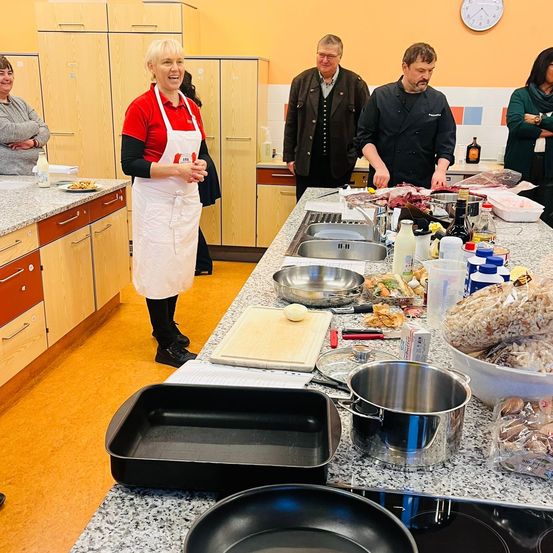 Eine Frau in einem roten Shirt und einer weißen Schürze steht in einer Küche und lacht. Sie ist von drei Männern in Kochuniformen umgeben. Die Arbeitsplatte ist mit Lebensmitteln und Kochutensilien übersät.