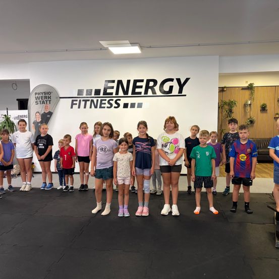 Eine Gruppe von Kindern und Jugendlichen steht vor einer Wand mit dem Wort 'ENERGY FITNESS' darauf. Sie tragen alle verschiedene farbige T-Shirts und Turnschuhe.