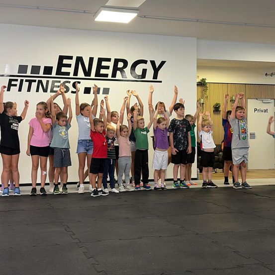Eine Gruppe von Kindern in Sportbekleidung steht in einer Reihe und hebt ihre Hände in einem Fitnessstudio mit einer großen Wand, auf der 'ENERGY FITNESS' steht.