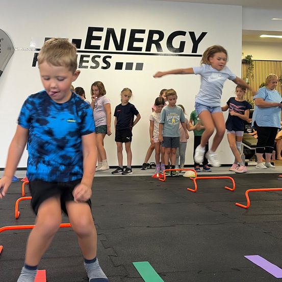 Kinder in einem Fitnessstudio, eines springt über Hürden, ein anderes geht. Eine Wand im Hintergrund sagt 'ENERGY FITNESS'.