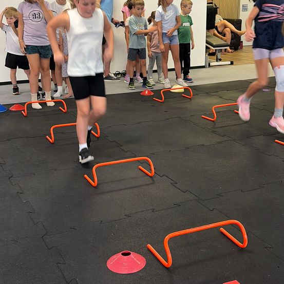 Eine Gruppe von Kindern nimmt an einer körperlichen Aktivität teil und läuft durch orange Hürden auf einem Fitnessstudioboden.