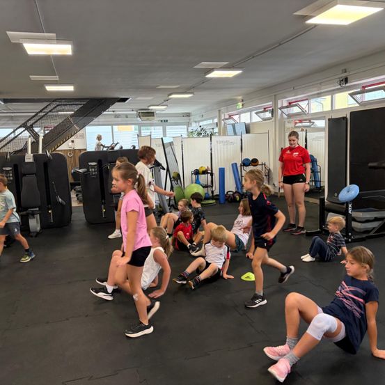 Eine Gruppe von Kindern nimmt an einem Fitnesskurs in einem Fitnessstudio teil. Einige sitzen auf dem Boden, während andere stehen und gehen. Eine Frau in einem roten Shirt beaufsichtigt.