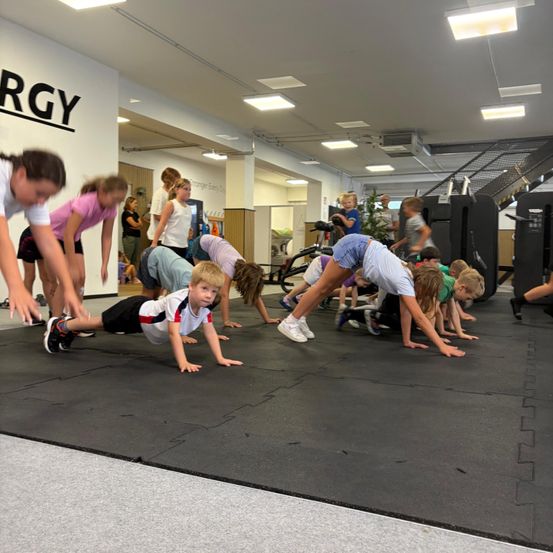 Eine Gruppe von Kindern macht Liegestütze auf einer Matte in einem Fitnessstudio, mit einer Wand im Hintergrund, auf der 'ergy' steht.