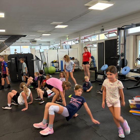 Eine Gruppe von Kindern trainiert im Fitnessstudio. Einige machen Dehnübungen, während andere gehen. Eine Frau in einem roten Shirt steht im Hintergrund.