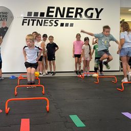 A group of children are participating in a fitness activity at a gym, jumping over orange hurdles on a black floor.