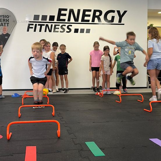 Eine Gruppe von Kindern nimmt in einem Fitnessstudio an einer Fitnessaktivität teil und springt über orange Hürden auf einem schwarzen Boden.