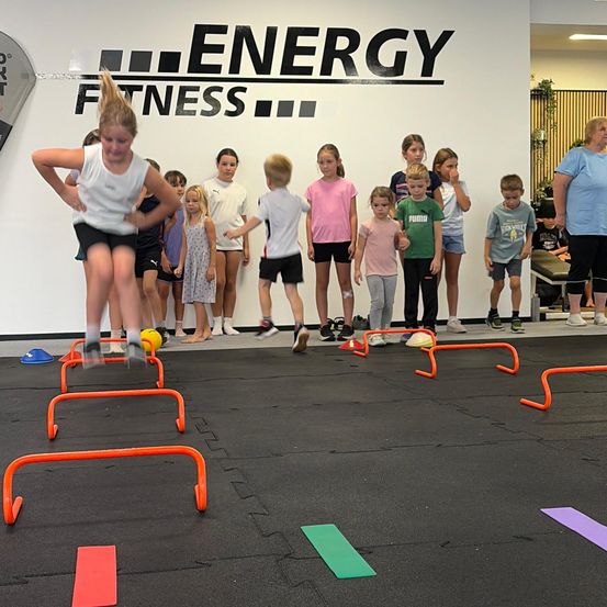 Eine Gruppe von Kindern nimmt an einer Fitnessaktivität teil, springt über orange Hürden auf einem schwarzen Boden, mit einer Wand im Hintergrund, auf der 'ENERGY FITNESS' steht.