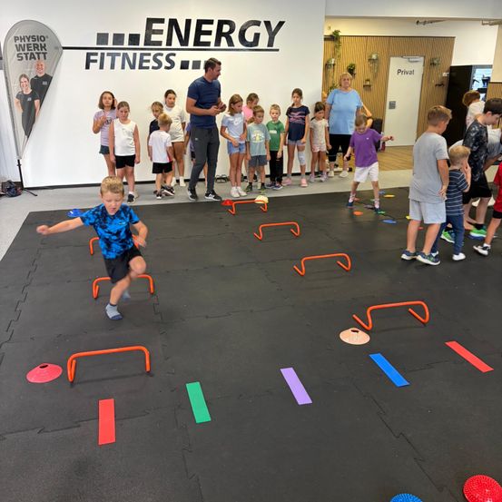 Eine Gruppe von Kindern nimmt in einem Fitnessstudio an einem Hindernisparcours teil, wobei ein Mann sie anleitet. Der Boden ist mit bunten Matten und Hindernissen bedeckt.