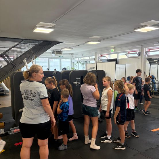 Eine Gruppe von Kindern und einer Frau steht in einem Fitnessstudio und hört einer Frau zu, die sie anweist. Das Fitnessstudio hat einen schwarzen Boden und verschiedene Fitnessgeräte im Hintergrund.