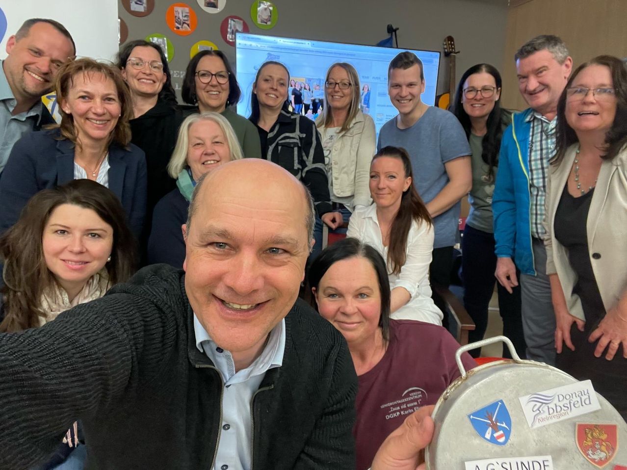 Gruppe von Menschen, die lächelnd in einem Raum stehen, mit einem Mann, der ein Selfie macht, einem Bildschirm, der Bilder hinter ihnen zeigt, und einer Frau, die ein Metallobjekt mit AG SUNDE Logo hält.