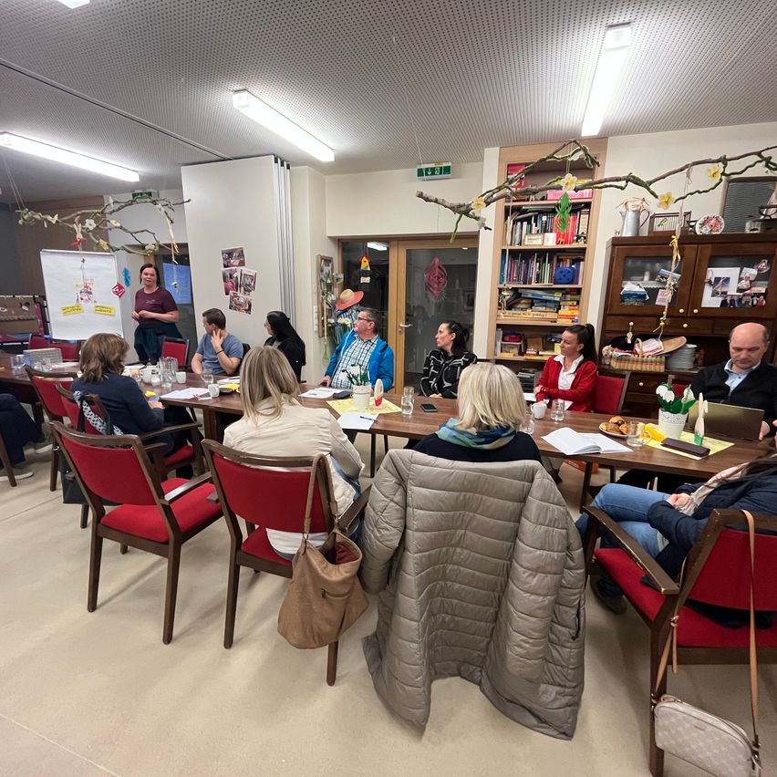 Gruppe von Erwachsenen sitzt um einen Tisch mit Papieren und einer Tafel in einem Raum.