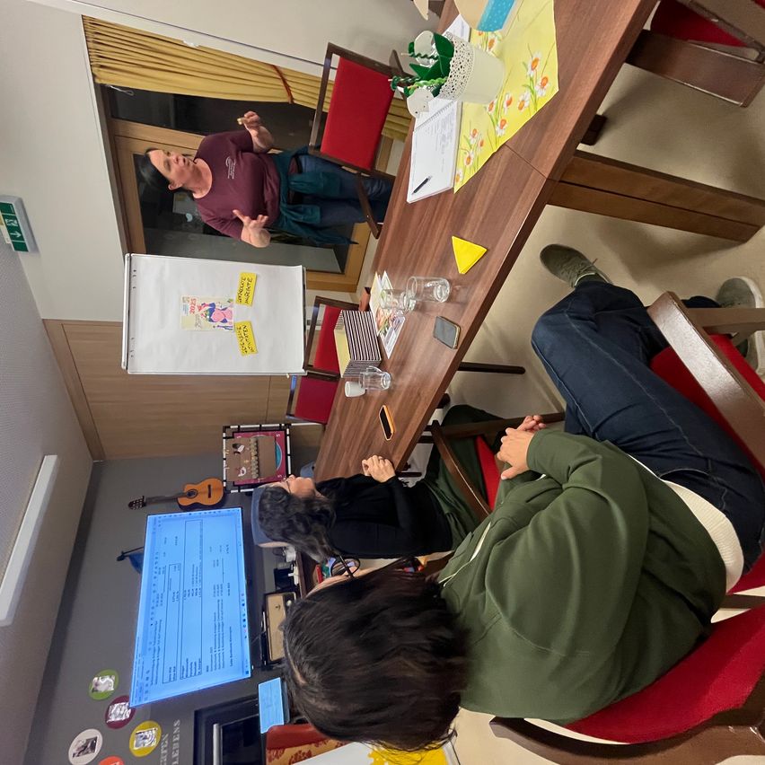 Ein Klassenzimmer mit einem Lehrer, der vor einer Tafel steht, und drei Schülern, die an einem Tisch sitzen. Ein Schüler schaut auf den Bildschirm eines Computers, während ein anderer sich auf den Tisch stützt. Ein gelbes Objekt und eine Gitarre hängen an der Wand. Ein roter Stuhl steht neben dem Tisch.