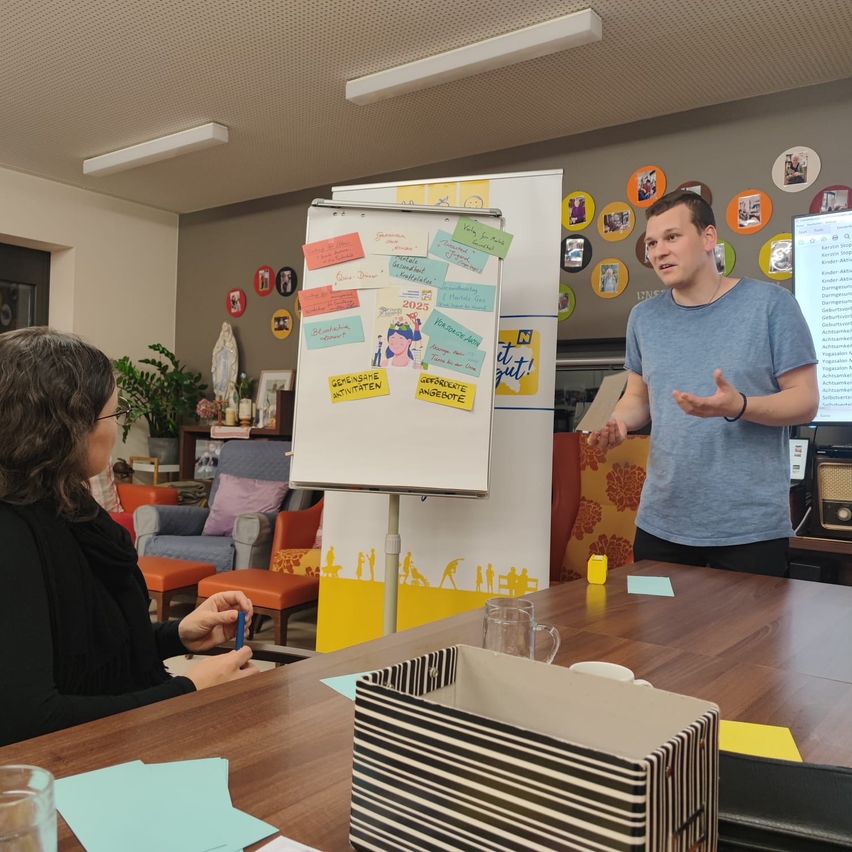 Ein Mann in einem blauen T-Shirt steht und gestikuliert, während er vor einem Tisch spricht. Eine Frau in einem schwarzen T-Shirt sitzt und schaut ihn an. Der Tisch hat eine Aufbewahrungsbox, ein Glas und Papiere. Eine Whiteboard ist an einem Ständer mit Haftnotizen und einem gelben Rand befestigt.